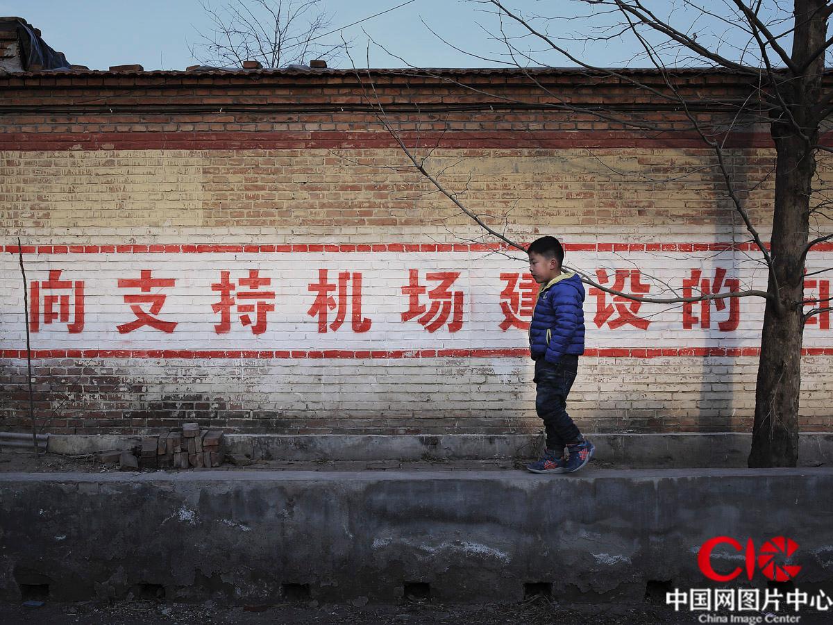 2016年2月，七岁的王琰浩从机场建设的宣传标语前走过，不久他将和父母一起搬离这个世代居住的村庄。